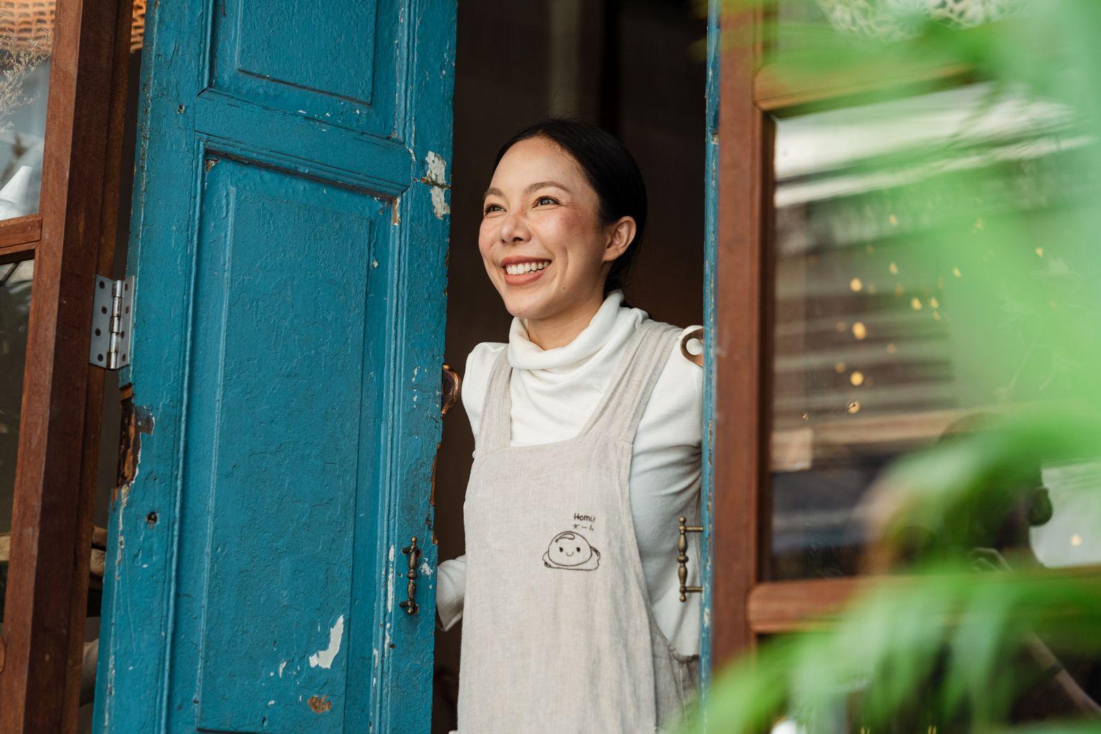 Smiling Asian café owner in an apron standing at the doorway of her small shop